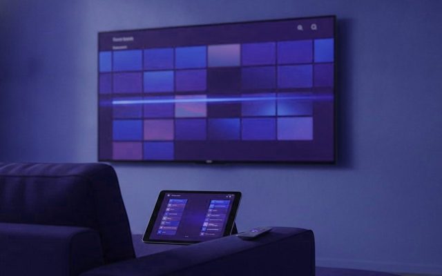 A TV showing a programme guide grid with a tablet and phone showing channel lists on a sofa